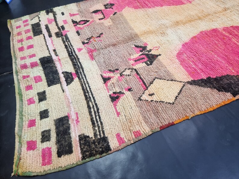 May include: A vintage Moroccan rug with a geometric pattern in shades of pink, black, and white. The rug has a distressed look and is made of wool.