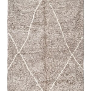 May include: A rectangular area rug with a thick, shaggy pile in a muted taupe colour. The rug features a large, white, diamond-shaped geometric pattern outlined in thin lines. The top and bottom edges are finished with short, white fringe tassels.
