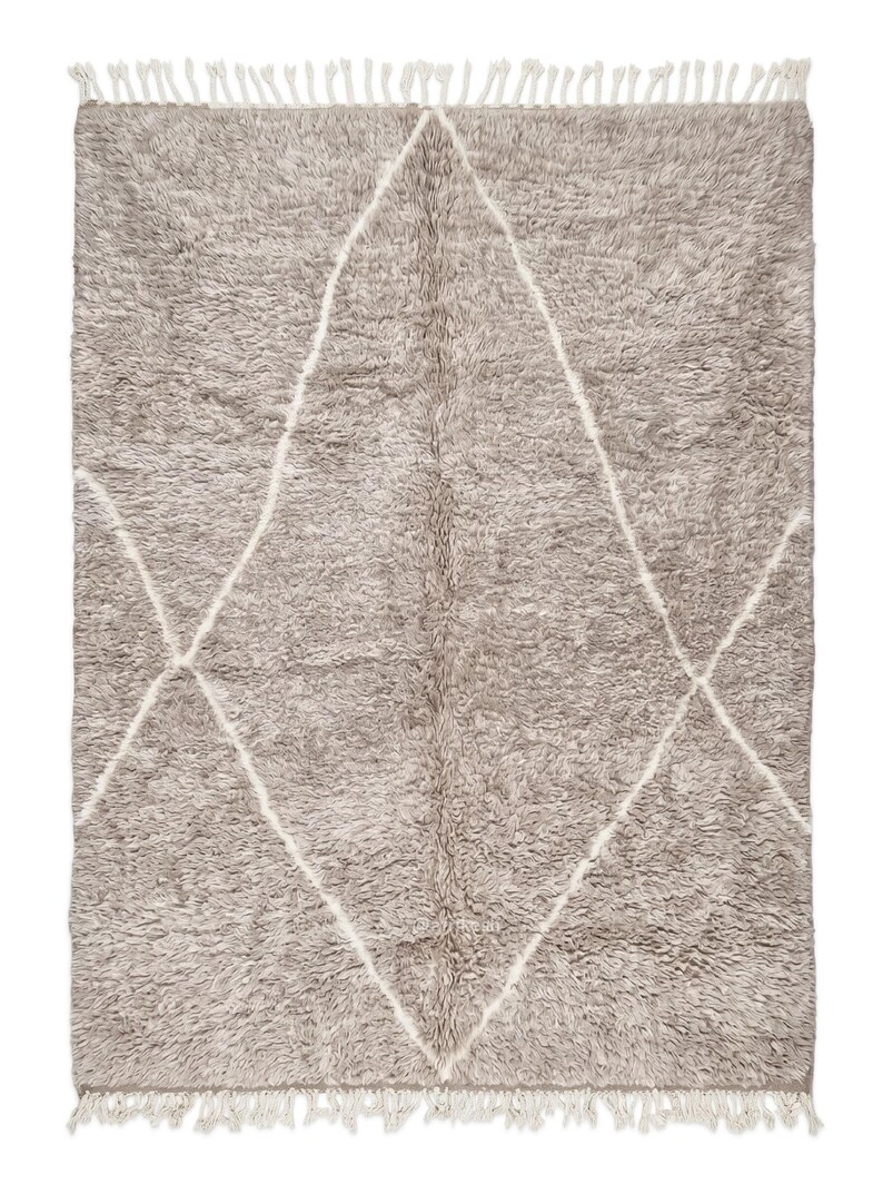 May include: A rectangular area rug with a thick, shaggy pile in a muted taupe colour. The rug features a large, white, diamond-shaped geometric pattern outlined in thin lines. The top and bottom edges are finished with short, white fringe tassels.