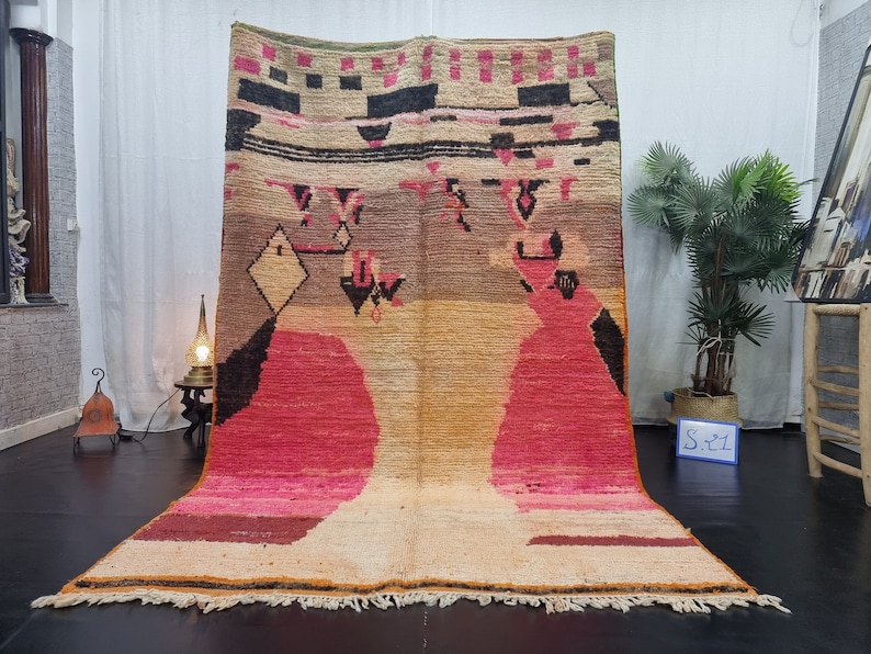 May include: A large, hand-woven rug with a geometric pattern in shades of pink, orange, brown, and black. The rug has a fringe border and is made of wool.