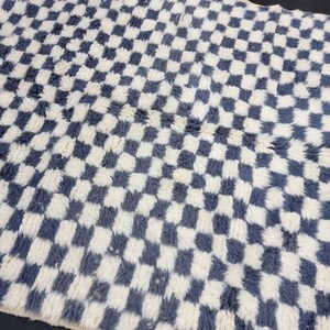 Op de afbeelding: Een wit en grijs geruit tapijt met een pluche textuur. Het tapijt is gemaakt van een zacht, pluche materiaal en heeft een modern, geometrisch ontwerp.