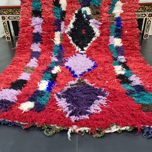 May include: A colorful Moroccan rug with a red background and a diamond pattern. The rug is made of wool and has a shaggy texture.