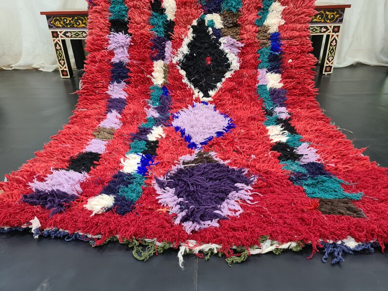 May include: A colorful Moroccan rug with a red background and a diamond pattern. The rug is made of wool and has a shaggy texture.