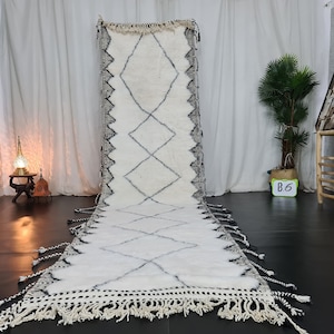 May include: A white and black Moroccan rug with a diamond pattern and fringe. The rug is laid out on a dark wood floor.