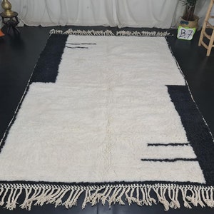 May include: A white and black geometric patterned rug with fringe on all sides. The rug has a modern design with black lines on a white background.