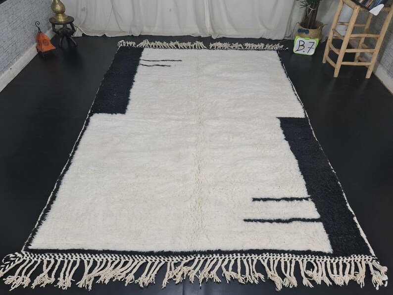 May include: A white and black geometric patterned rug with fringe on all sides. The rug has a modern design with black lines on a white background.