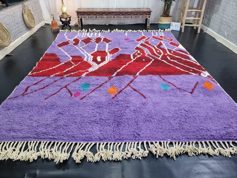 May include: A purple and red Moroccan rug with a geometric design and white fringe. The rug has a variety of colorful accents, including turquoise, pink, and orange.