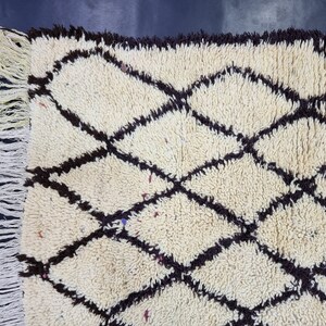 May include: A white and brown Moroccan rug with a diamond pattern. The rug has a fringe border.