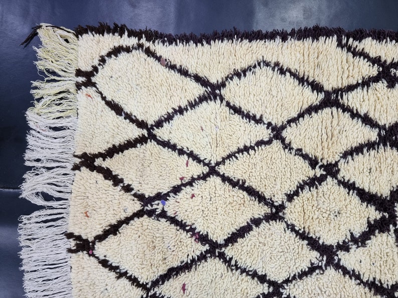 May include: A white and brown Moroccan rug with a diamond pattern. The rug has a fringe border.