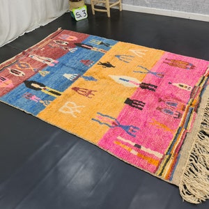 May include: A colorful Moroccan rug with a geometric pattern and fringe. The rug is made of wool and has a variety of colors, including red, blue, yellow, and pink.