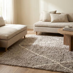 May include: A rectangular area rug with a thick, shaggy, light brown pile featuring a white geometric diamond pattern and fringed edges. The rug is positioned on a wooden floor in front of a beige sofa, a matching chaise lounge, and a round wooden table.