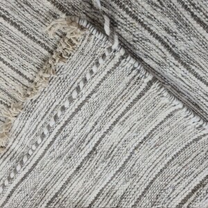 Puede incluir: Primer plano de una alfombra tejida blanca y gris con borde de flecos. La alfombra tiene un sutil patr&oacute;n de rayas.
