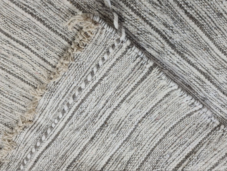 Puede incluir: Primer plano de una alfombra tejida blanca y gris con borde de flecos. La alfombra tiene un sutil patr&oacute;n de rayas.