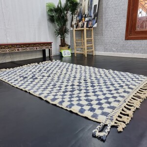 Op de afbeelding: Een wit en blauw geruit tapijt met franjes aan de randen. Het tapijt is gemaakt van wol en heeft een zachte, pluche textuur.