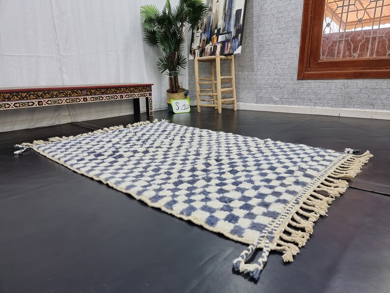Op de afbeelding: Een wit en blauw geruit tapijt met franjes aan de randen. Het tapijt is gemaakt van wol en heeft een zachte, pluche textuur.
