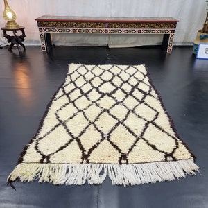 May include: A white and brown Moroccan rug with a diamond pattern and fringe. The rug is laid out on a black floor.