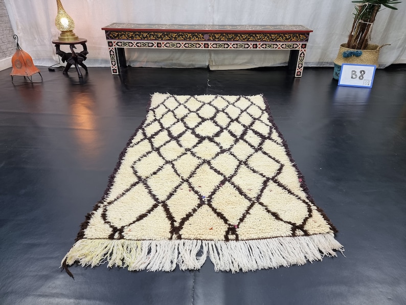 May include: A white and brown Moroccan rug with a diamond pattern and fringe. The rug is laid out on a black floor.