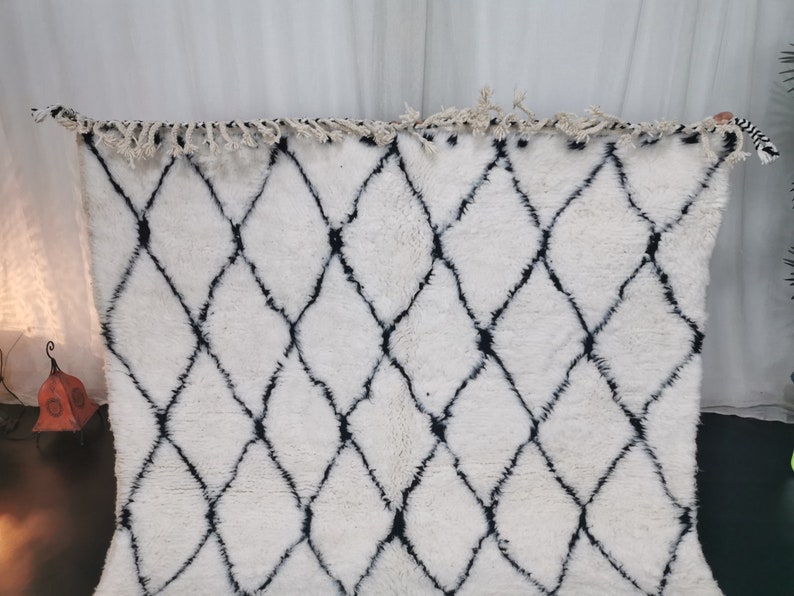 May include: White and black Moroccan rug with a diamond pattern. The rug has a fringe border.