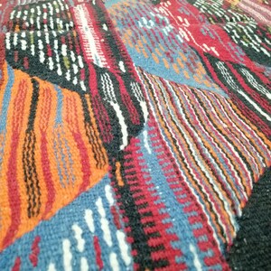 Pu&ograve; includere: Primo piano di un tessuto intrecciato con un motivo geometrico. Il design presenta un patchwork di forme in rosso, arancione, blu, nero e bianco. La trama appare spessa e strutturata, suggerendo un tappeto o una coperta.