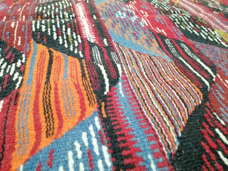 Pu&ograve; includere: Primo piano di un tessuto intrecciato con un motivo geometrico. Il design presenta un patchwork di forme in rosso, arancione, blu, nero e bianco. La trama appare spessa e strutturata, suggerendo un tappeto o una coperta.
