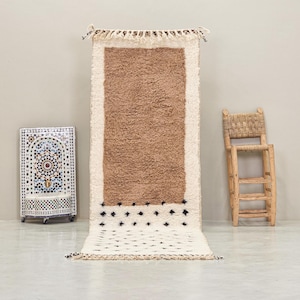 Op de afbeelding: Een rechthoekig, crèmekleurig tapijt met een bruin midden en een crèmekleurige rand. Het tapijt heeft een textuur en is versierd met zwarte ruitvormen. Een kleine houten stoel en een decoratieve mozaïekfontein staan op de achtergrond.