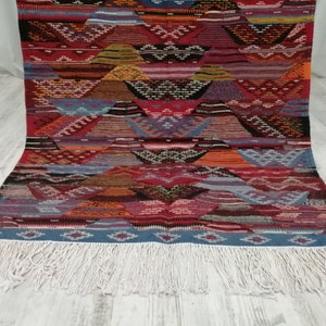 Pu&ograve; includere: Un tappeto colorato con motivi geometrici e bordo con frange. Il tappeto presenta un motivo ripetuto di forme triangolari nei toni del rosso, blu, arancione e marrone. La frangia &egrave; bianca.