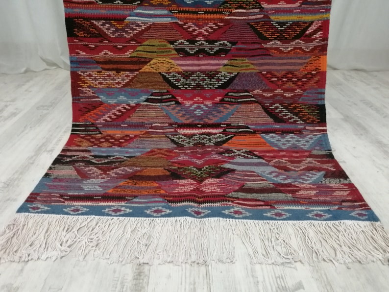 Pu&ograve; includere: Un tappeto colorato con motivi geometrici e bordo con frange. Il tappeto presenta un motivo ripetuto di forme triangolari nei toni del rosso, blu, arancione e marrone. La frangia &egrave; bianca.