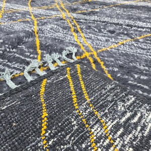 May include: Close-up of a gray and white woven rug with yellow stripes. The rug has a fringe border.