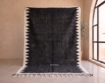 May include: A large rectangular area rug featuring a solid black centre with a distressed texture. The rug is bordered by a wide white band with a repeating black sawtooth pattern along the edges. The bottom edge has white fringe tassels.