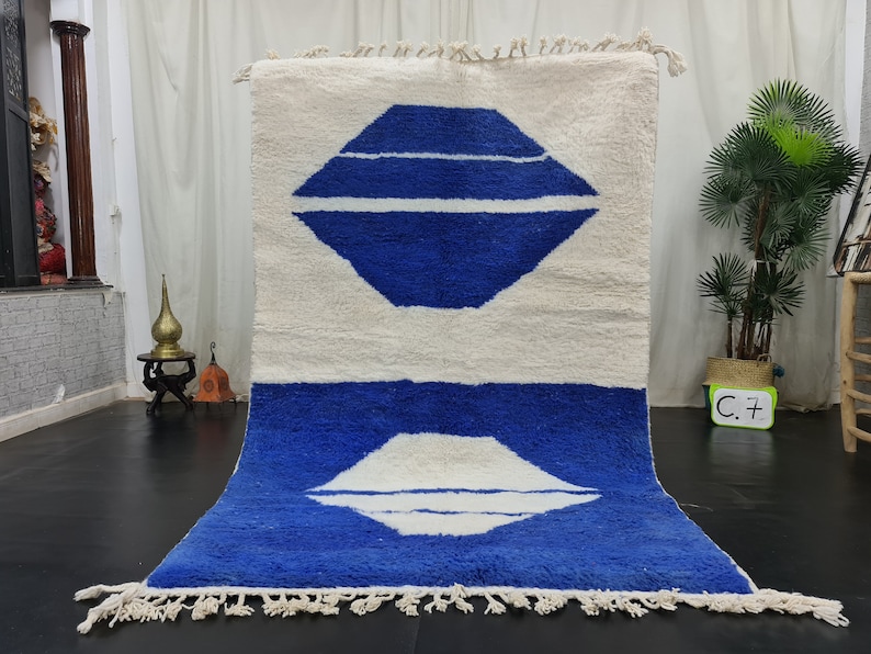 Puede incluir: Una alfombra marroqu&iacute; blanca y azul con un dise&ntilde;o geom&eacute;trico. La alfombra tiene un fondo blanco con dos formas de diamantes azules, cada una con una franja blanca en el centro. La alfombra tiene un borde de flecos.