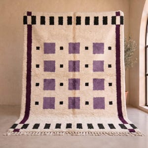 Puede incluir: Una alfombra rectangular con base de color crema que presenta una cuadrícula de cuadrados morados y pequeños cuadrados negros. La alfombra está enmarcada por un borde morado grueso y un patrón de rectángulos negros y crema alternos con flecos.