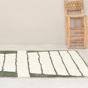 May include: A rectangular rug with a white background and green rectangular shapes. The rug has a fringed edge and is placed on a light-colored floor. A wooden chair with a woven back is in the background.