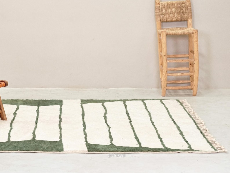 May include: A rectangular rug with a white background and green rectangular shapes. The rug has a fringed edge and is placed on a light-colored floor. A wooden chair with a woven back is in the background.
