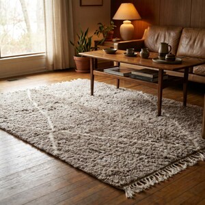 May include: A rectangular, high-pile area rug in a neutral beige tone, featuring a subtle white diamond-patterned design. The rug has a thick, textured surface and is finished with short, white fringe detailing along the shorter edges.