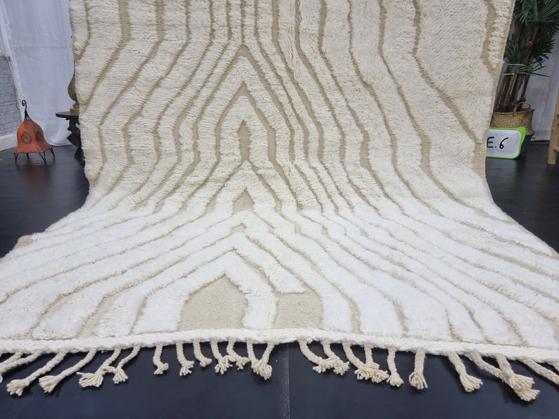 May include: A white and cream colored rug with a geometric pattern. The rug has a fringe border.
