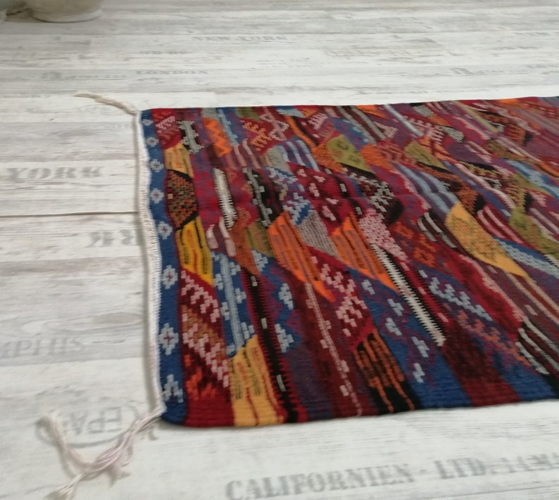 Pu&ograve; includere: Un tappeto rettangolare colorato con un design patchwork. Il tappeto presenta una variet&agrave; di colori tra cui rosso, blu, arancione e giallo. I bordi sono rifiniti con un bordo blu e frange bianche.