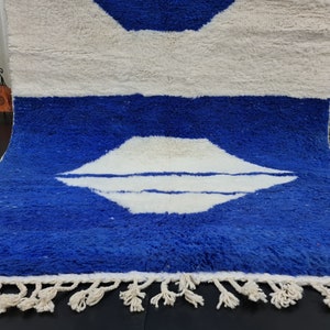Puede incluir: Una alfombra marroqu&iacute; blanca y azul con un patr&oacute;n geom&eacute;trico. La alfombra tiene un borde de flecos y est&aacute; hecha de lana.