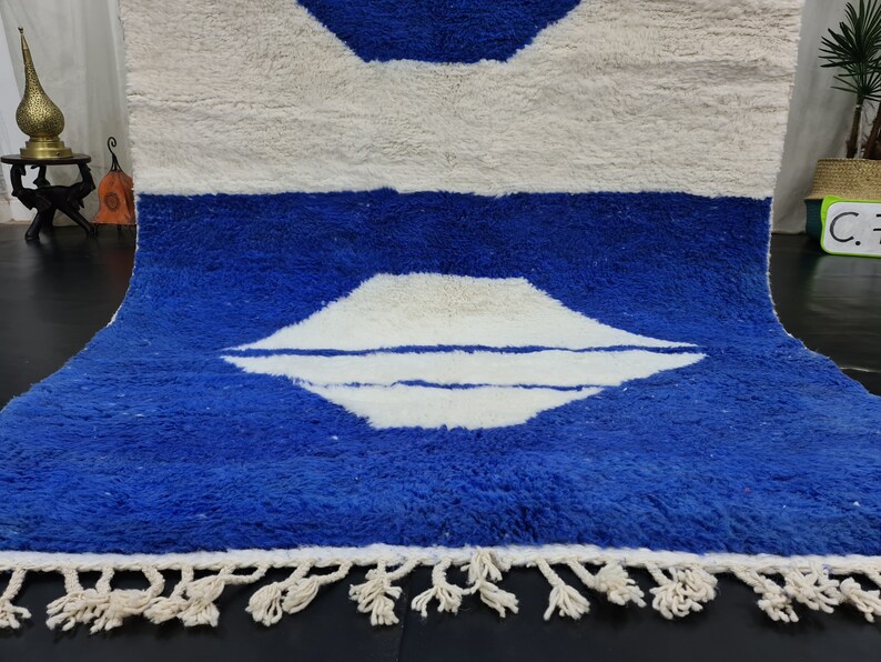 Puede incluir: Una alfombra marroqu&iacute; blanca y azul con un patr&oacute;n geom&eacute;trico. La alfombra tiene un borde de flecos y est&aacute; hecha de lana.
