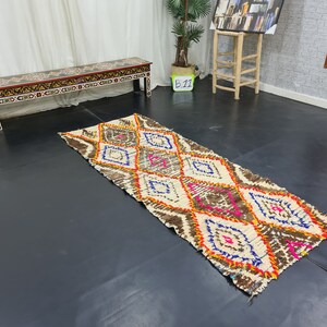 May include: A white and brown patterned rug with pink, orange, and blue accents. The rug has a diamond pattern and is laid out on a dark floor.
