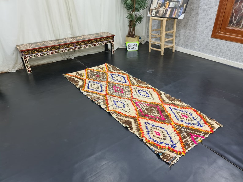 May include: A white and brown patterned rug with pink, orange, and blue accents. The rug has a diamond pattern and is laid out on a dark floor.