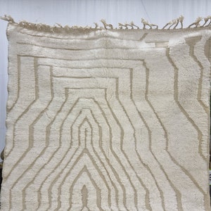 May include: A white Moroccan rug with a geometric pattern in a lighter shade of beige. The rug has a fringe border.