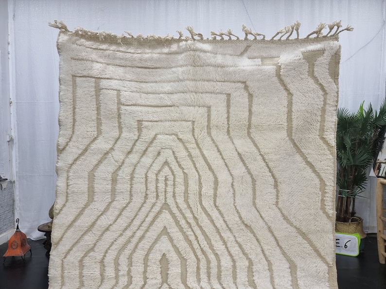 May include: A white Moroccan rug with a geometric pattern in a lighter shade of beige. The rug has a fringe border.