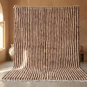 Peut inclure: Un grand tapis rectangulaire avec un fond crème et des rayures verticales marron. Le tapis est fait d'une matière épaisse et texturée et a un bord effiloché. Le tapis est exposé dans une pièce avec de la lumière naturelle.