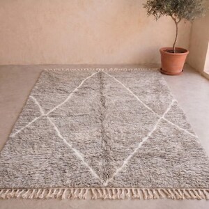 May include: A rectangular area rug featuring a thick, shaggy pile in a mottled grey colour. The rug is decorated with a large, white, hand-drawn diamond pattern that spans the centre. The short ends of the rug are finished with cream-coloured fringe.