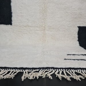May include: White and black wool rug with a geometric pattern and fringe. The rug has a black border and two black lines in the centre.