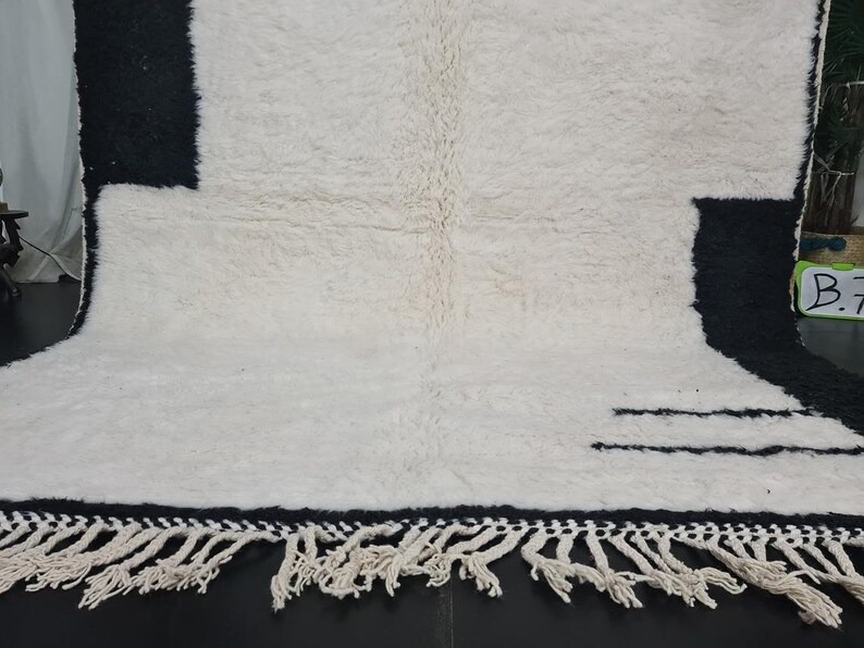 May include: White and black wool rug with a geometric pattern and fringe. The rug has a black border and two black lines in the centre.
