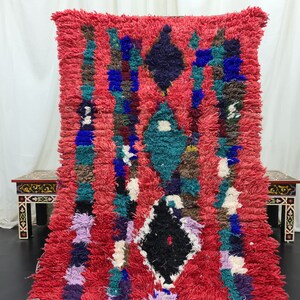 May include: A red, blue, green, and black Moroccan rug with a geometric pattern. The rug is made of wool and has a soft, fluffy texture.