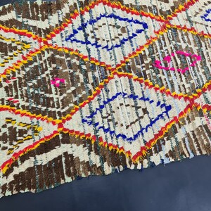 May include: A white, brown, blue, yellow, and pink Moroccan rug with a geometric pattern. The rug is handwoven and has a fringed edge.