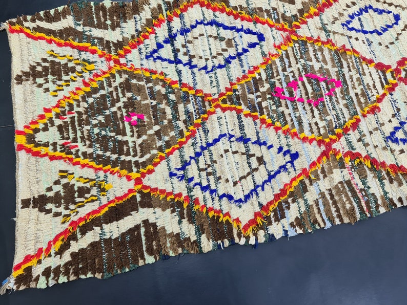 May include: A white, brown, blue, yellow, and pink Moroccan rug with a geometric pattern. The rug is handwoven and has a fringed edge.