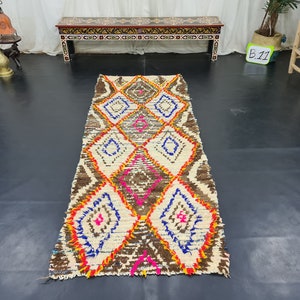 May include: A white, brown, orange, pink, and blue Moroccan rug with a diamond pattern. The rug is approximately 8 feet long and 2 feet wide.